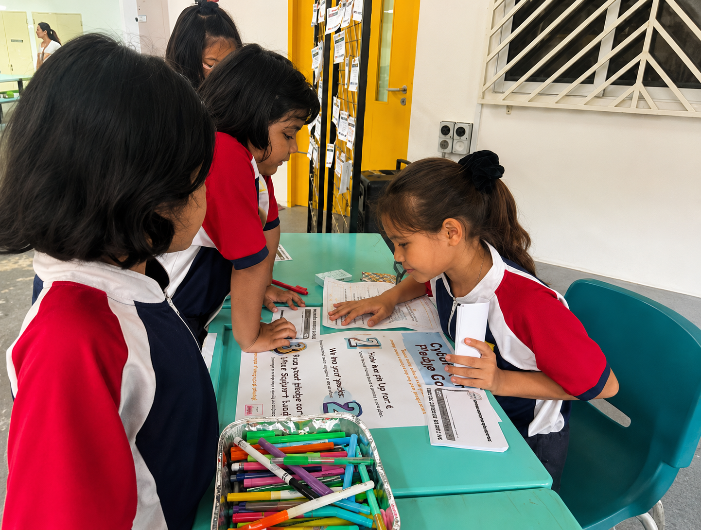 Three children in red and white uniforms lean over a table with papers and markers in a school setting.
